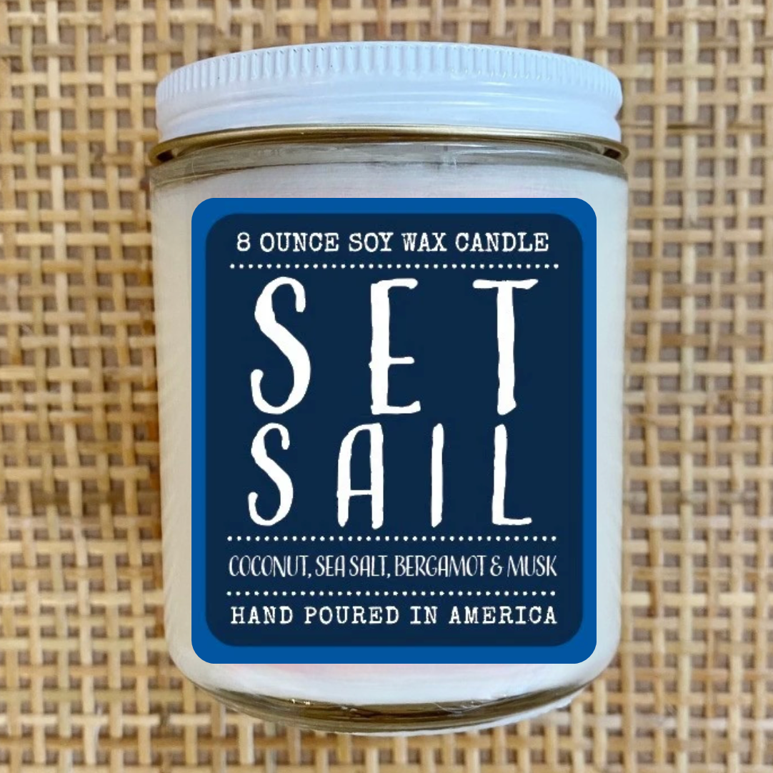 An 8-ounce soy wax candle in a clear glass jar with a white lid and a blue label reading 'SET SAIL'. The candle is centered against a woven tan background.