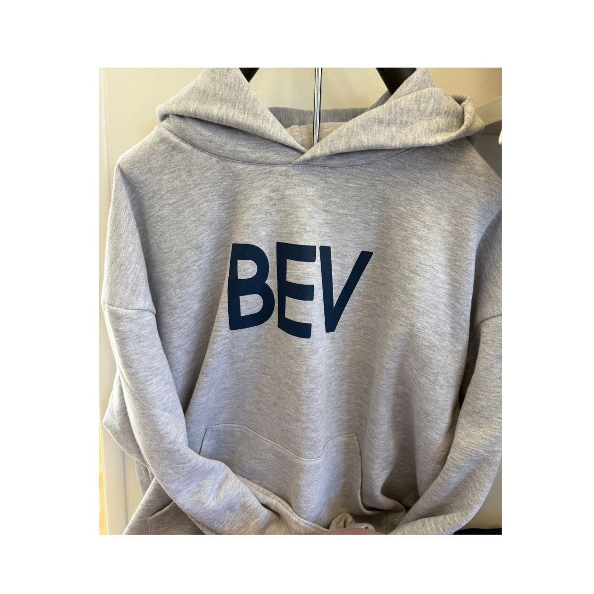 A light gray heather pullover hoodie with the letters 'BEV' printed in dark navy blue across the chest, hanging on a hanger against a neutral indoor background.