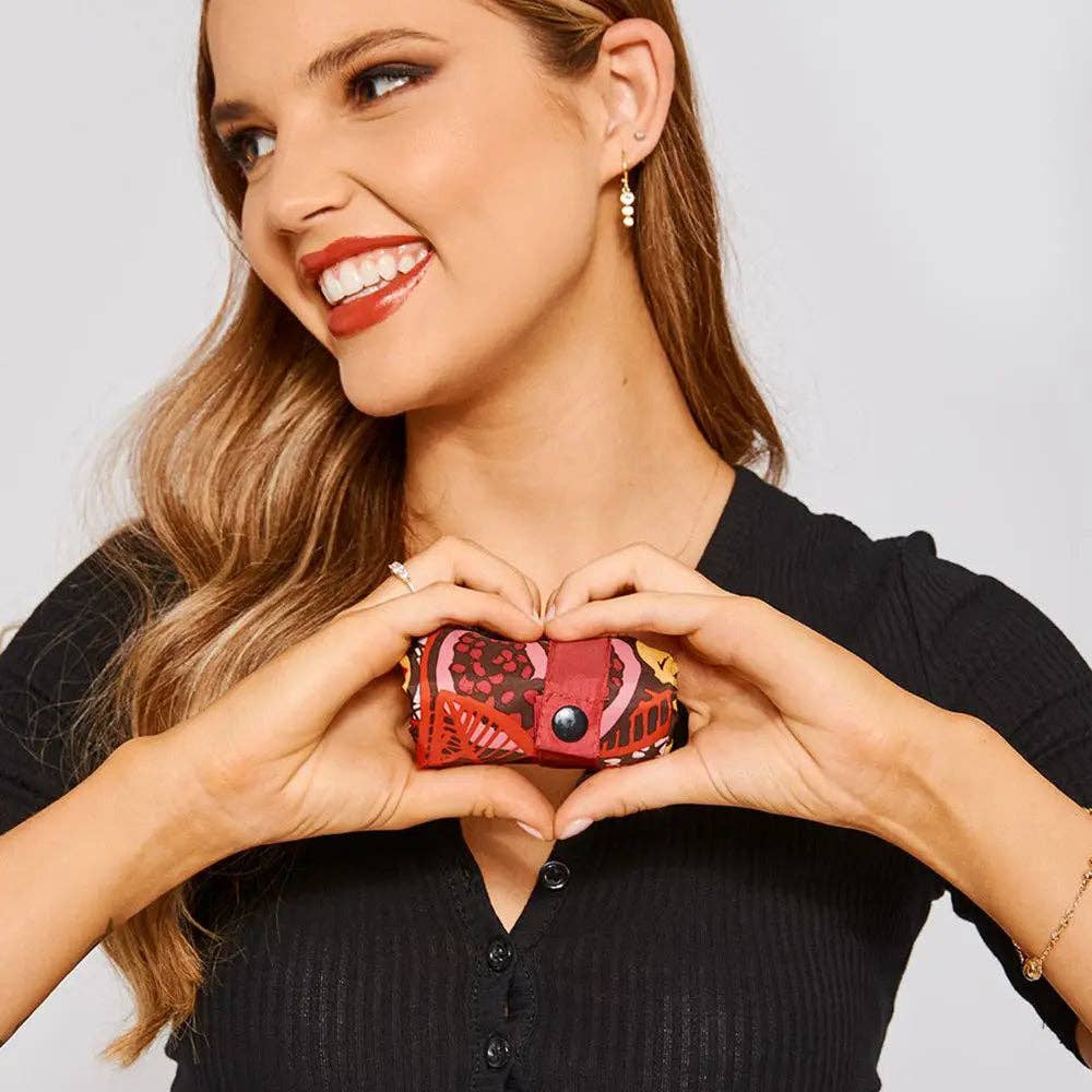 A woman in a black ribbed top holds a small red patterned clutch purse in front of her chest, forming a heart shape with her hands. The clutch has a red base with black and yellow abstract patterns and a silver snap button.