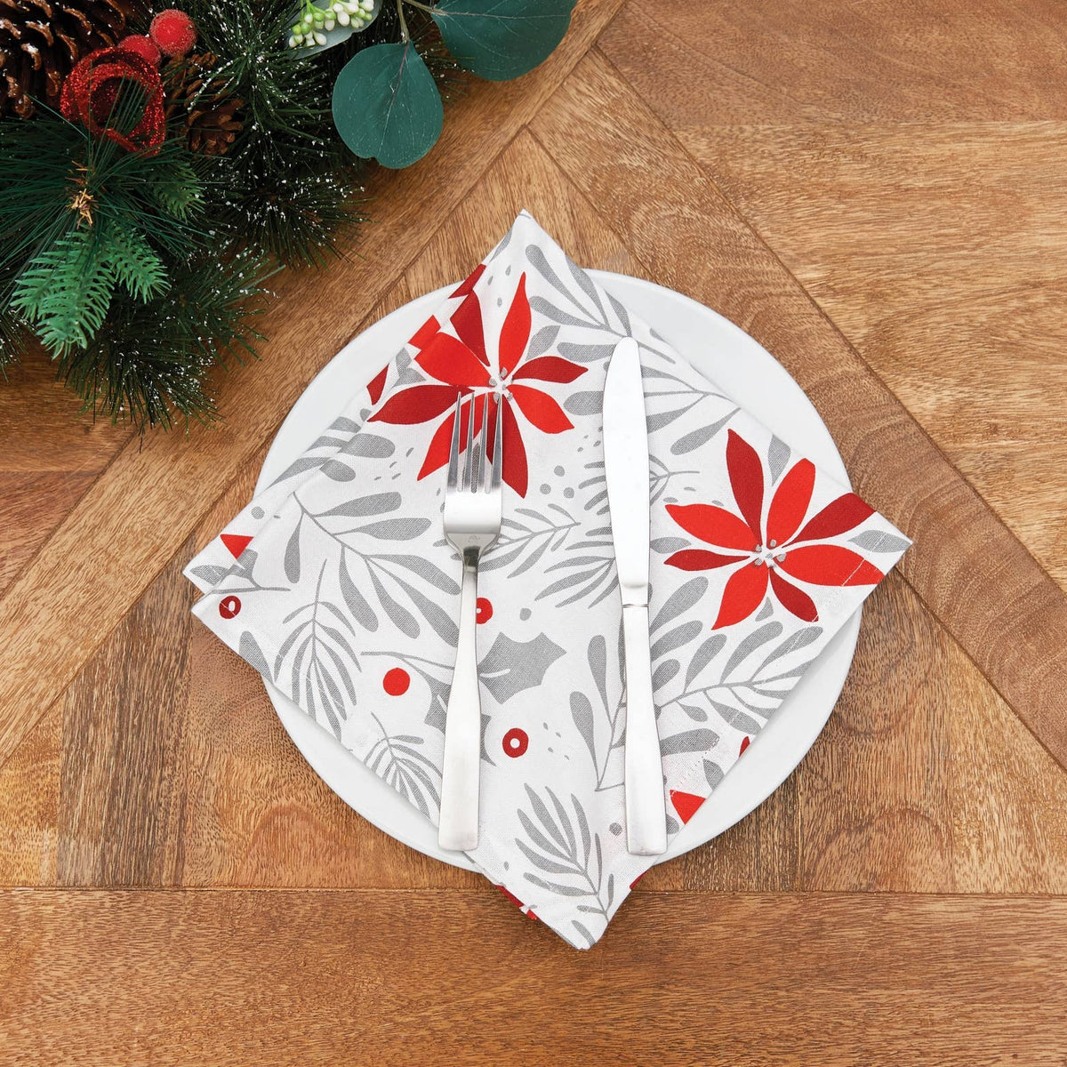 A festive holiday table setting featuring a white napkin with a red poinsettia and gray leaf pattern, folded around a fork and knife on a wooden table with holiday greenery in the corner.