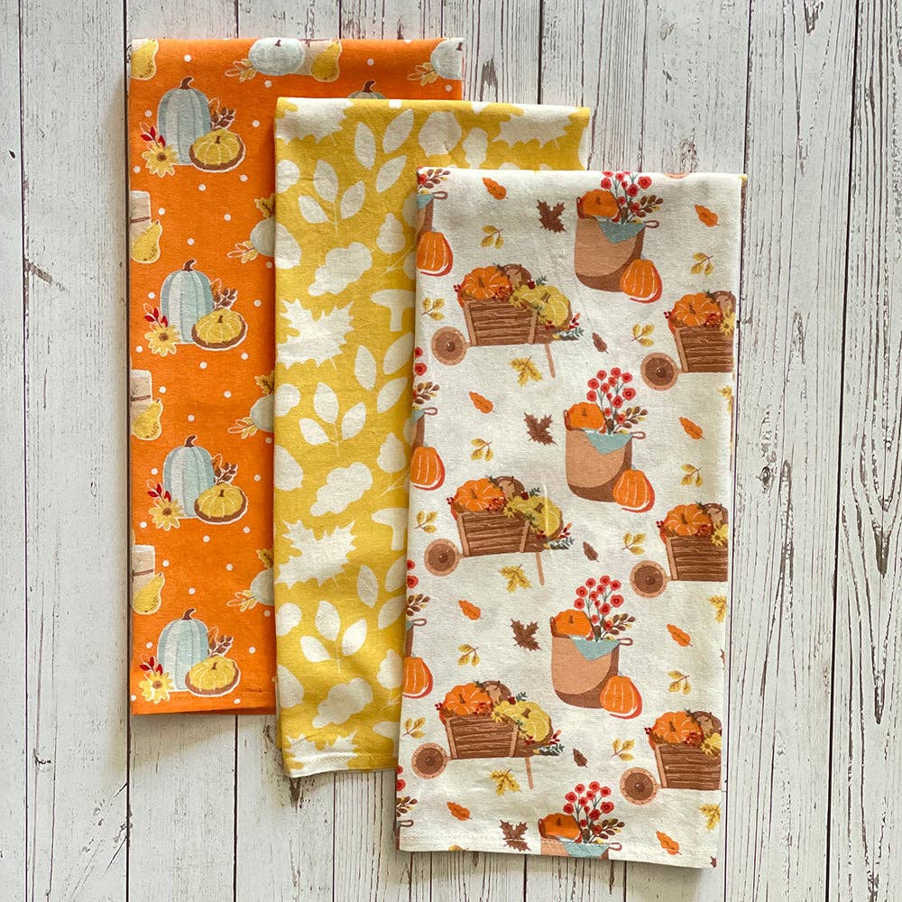 Three folded fabric napkins with autumn fall patterns are arranged on a white wood plank surface. The napkin on the left is orange with pumpkins, acorns, and leaves.