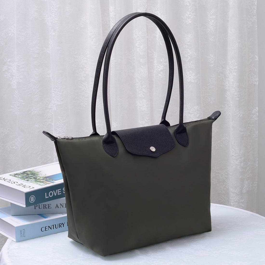 An olive green nylon tote bag with dark navy leather handles and a front flap, displayed on a white table next to a stack of books in front of a white curtain.