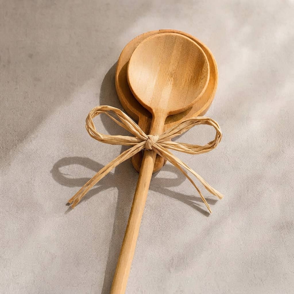 Two light natural wood spoons tied with a tan raffia ribbon bow, photographed on a textured light gray surface with soft shadows.