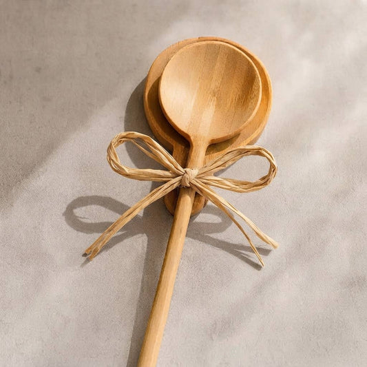 Two light natural wood spoons tied with a tan raffia ribbon bow, photographed on a textured light gray surface with soft shadows.