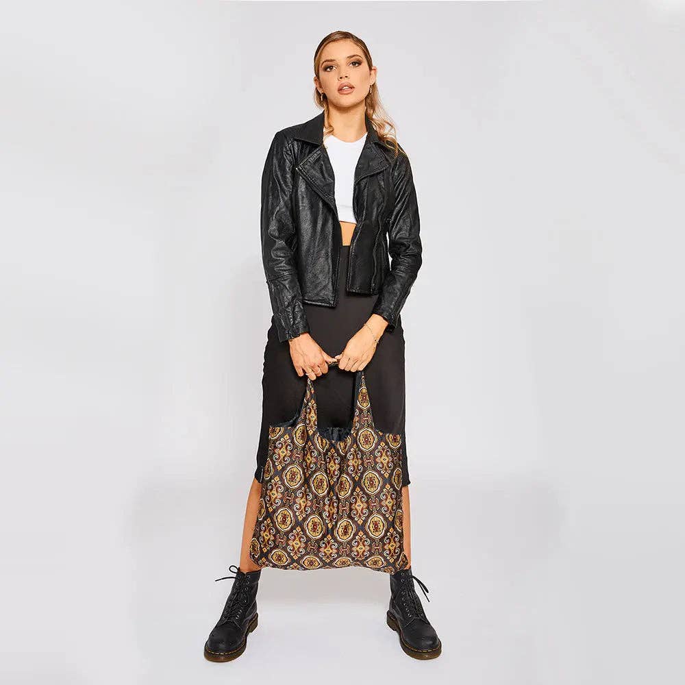 A model stands against a light gray studio background wearing a black faux-leather jacket, black skirt, white top, black boots, and holding a large black tote bag with a gold and brown geometric pattern.