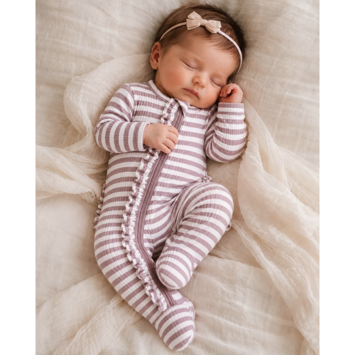 A sleeping newborn baby wearing a mauve and white striped footed sleeper with a ruffled zipper placket and a matching bow headband, lying on a soft cream-colored textured blanket.