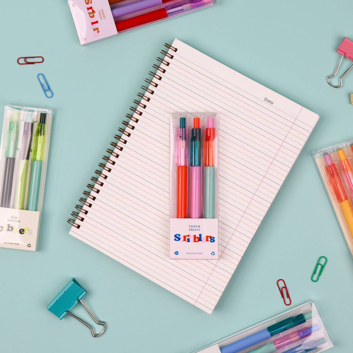 A flat-lay of a spiral-bound lined notebook centered on a light blue background, surrounded by stationery items including pens, highlighters, paper clips, and a package of Scribbl pens. The main product appears to be the notebook with the Scribbl pen pack attached.