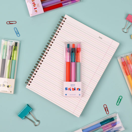 A flat-lay of a spiral-bound lined notebook centered on a light blue background, surrounded by stationery items including pens, highlighters, paper clips, and a package of Scribbl pens. The main product appears to be the notebook with the Scribbl pen pack attached.