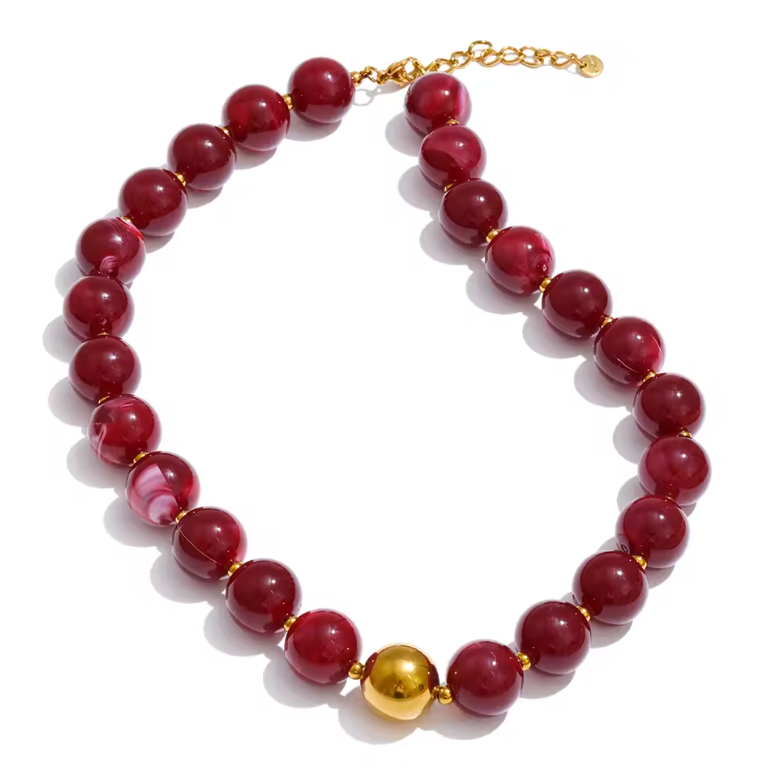 A beaded necklace made of glossy dark red round beads with small gold spacer rings and a single prominent gold sphere bead at the center, finished with a gold chain extender clasp on a white background.