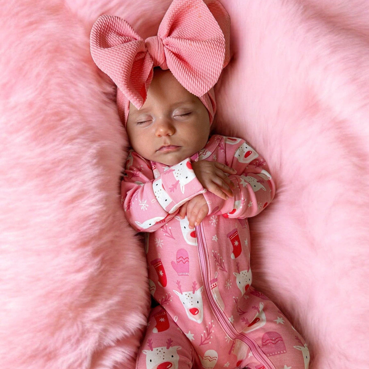 A sleeping newborn baby wearing a pink zip-up onesie with a playful cat and heart pattern, paired with a matching pink knit bow headband, lying on a fluffy pink blanket.
