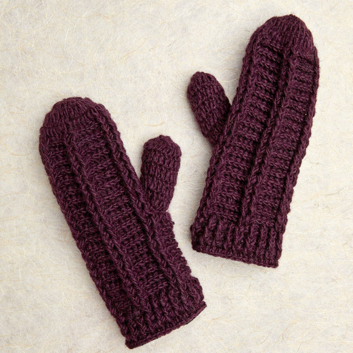 A pair of dark plum purple knit mittens laid flat on a textured off-white background.