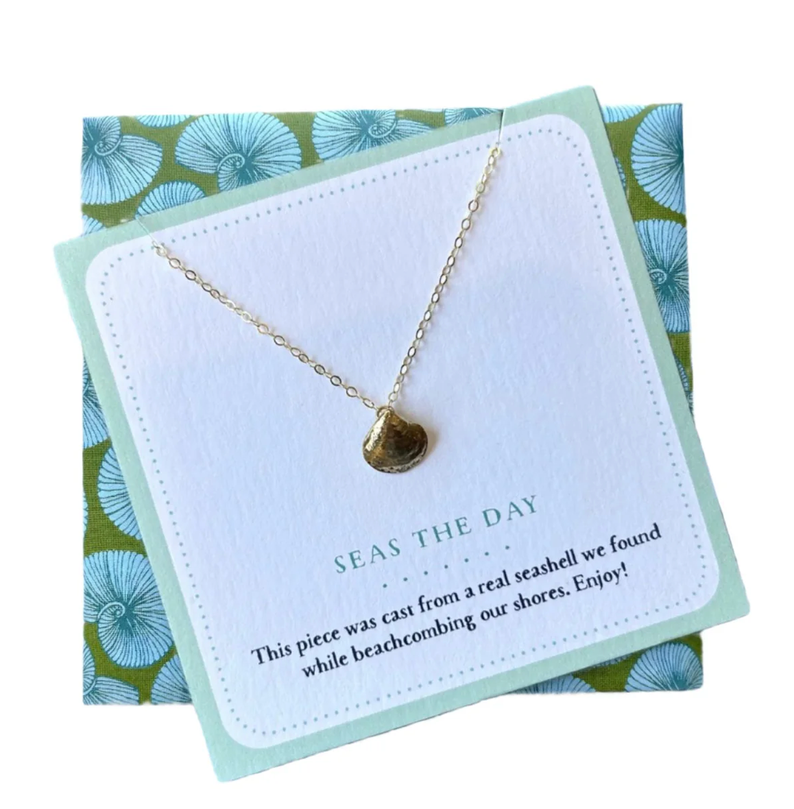 A gold necklace with a small seashell pendant displayed on a white card with a mint border, placed on top of a green and blue patterned gift box. The card reads 'SEAS THE DAY'.