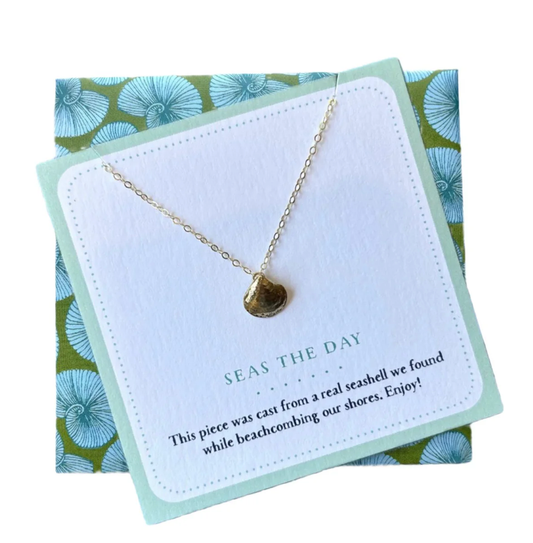A gold necklace with a small seashell pendant displayed on a white card with a mint border, placed on top of a green and blue patterned gift box. The card reads 'SEAS THE DAY'.