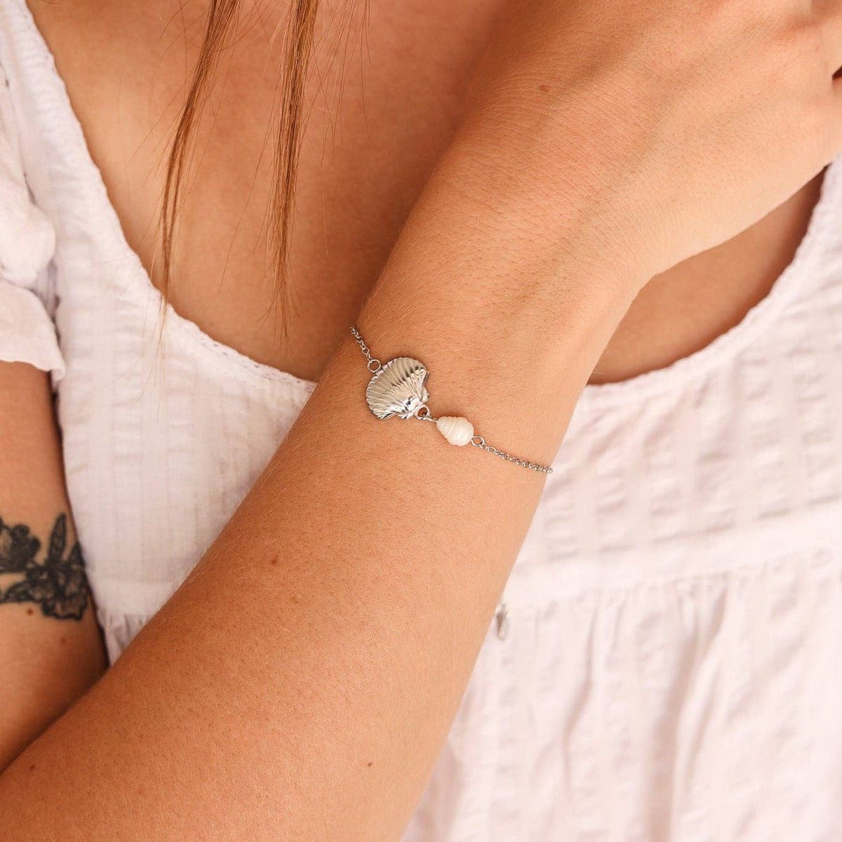 A close-up of a woman's wrist wearing a delicate silver bracelet with a small heart-shaped charm and a tiny white bead.