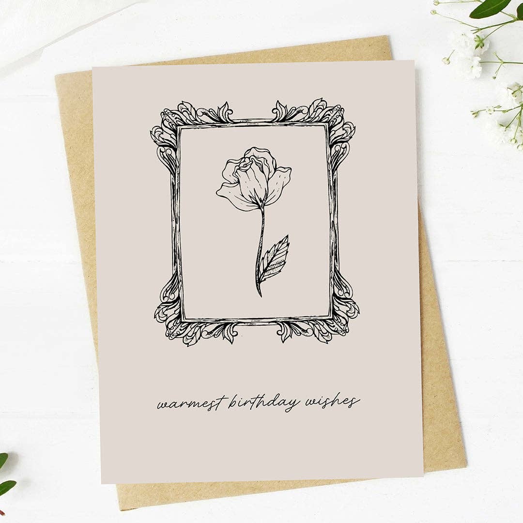 A square greeting card with a black line drawing of a rose inside an ornate frame, with the text 'warmest birthday wishes' at the bottom, shown on top of a kraft envelope on a white surface.