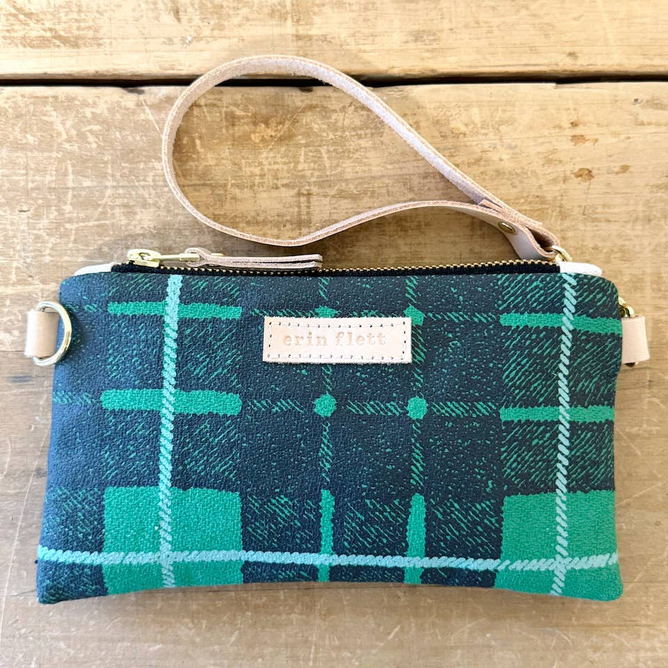 A small zip pouch with a green and navy plaid pattern, featuring a light beige wrist strap and a white brand label that reads 'erin flann'. The pouch is photographed flat on a rustic wooden surface.