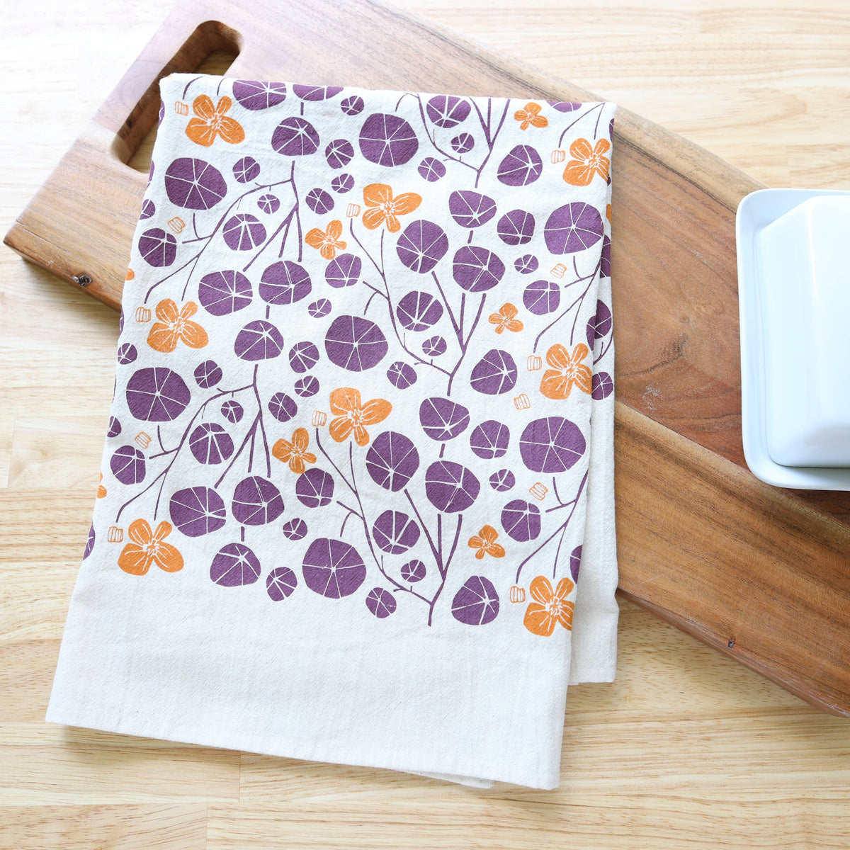 A white kitchen tea towel with a purple and orange floral pattern, styled on a wooden cutting board next to a white ceramic butter dish on a light wood tabletop.