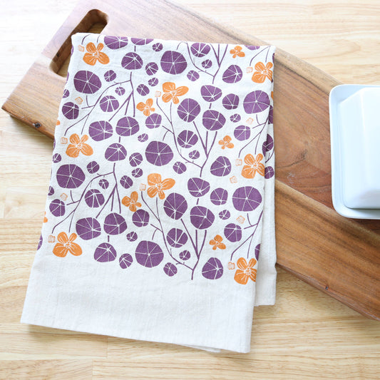 A white kitchen tea towel with a purple and orange floral pattern, styled on a wooden cutting board next to a white ceramic butter dish on a light wood tabletop.