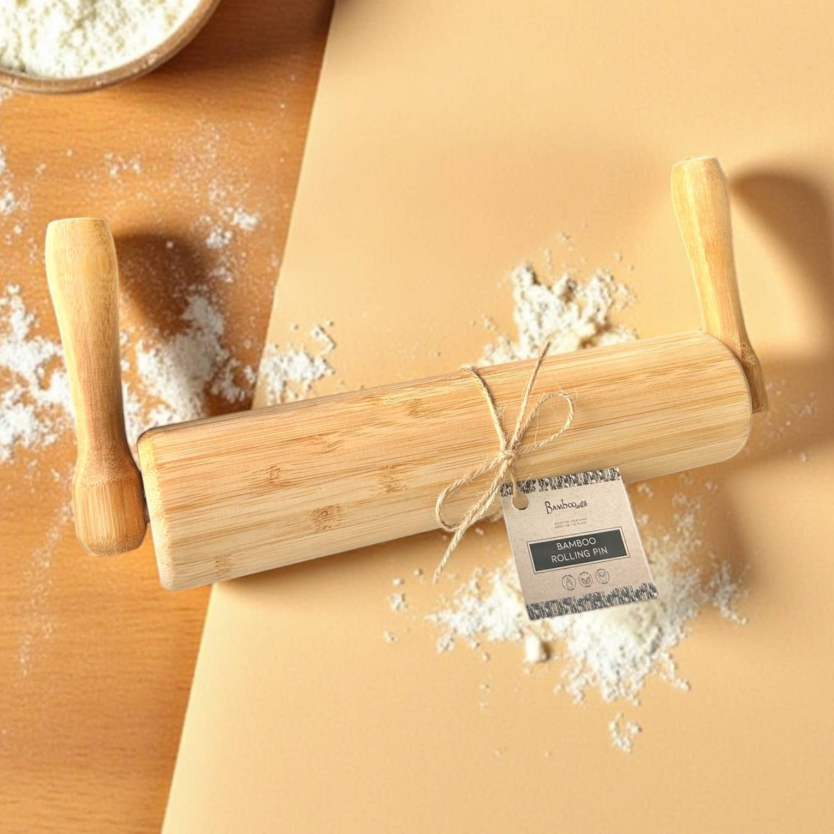 A bamboo rolling pin with wooden handles, tied with twine and a small paper tag, placed on a beige surface with scattered flour. A bowl of flour is partially visible in the top left corner.