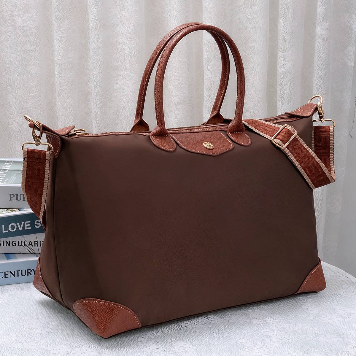 A brown nylon tote bag with brown leather handles, trim, and corner protectors, shown on a white surface with a white curtain background.