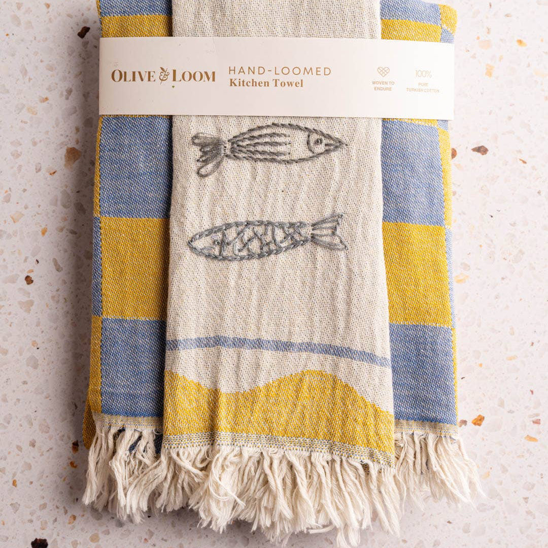 Hand-loomed kitchen towel with fish design and Olive & Loom branding on a textured surface.