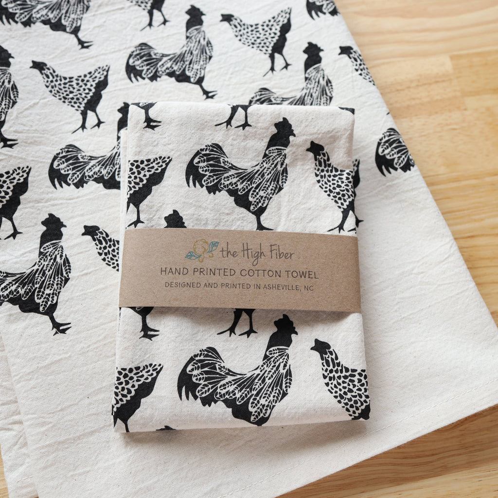 Folded hand-printed cotton towel with black rooster pattern on a wooden surface