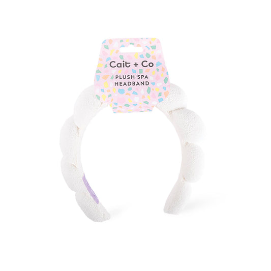 A white plush spa headband by Cait + Co, shown with its original pink and multicolor patterned cardboard hanger on a clean white background.