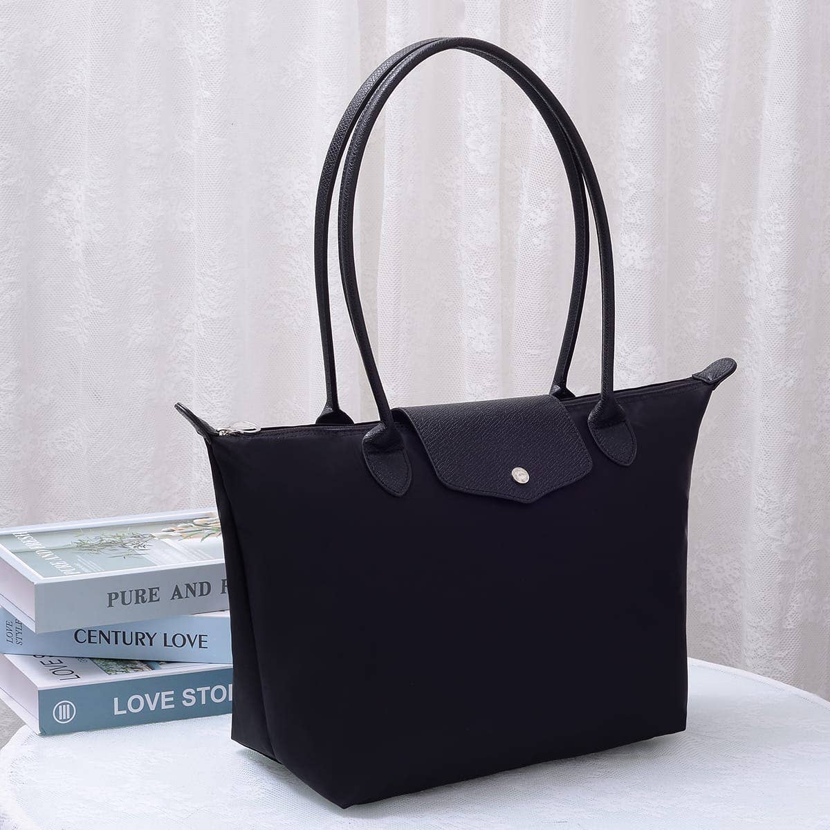 A black tote bag with double top handles and a front flap pocket, styled on a white surface next to stacked books against a white curtain backdrop.
