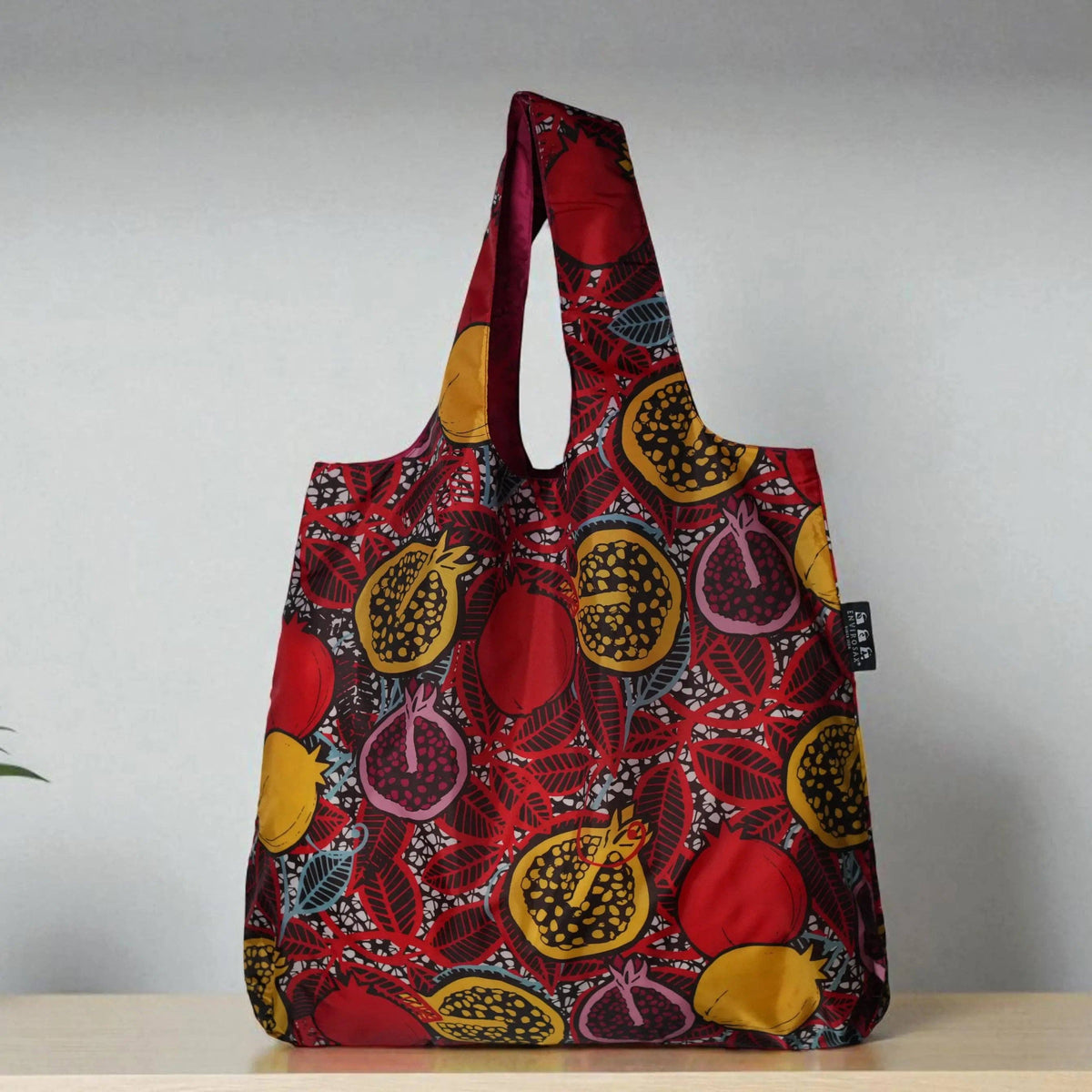 A reusable fabric tote bag with a bold red, yellow, and purple pomegranate print, standing on a light wooden surface against a neutral gray background.