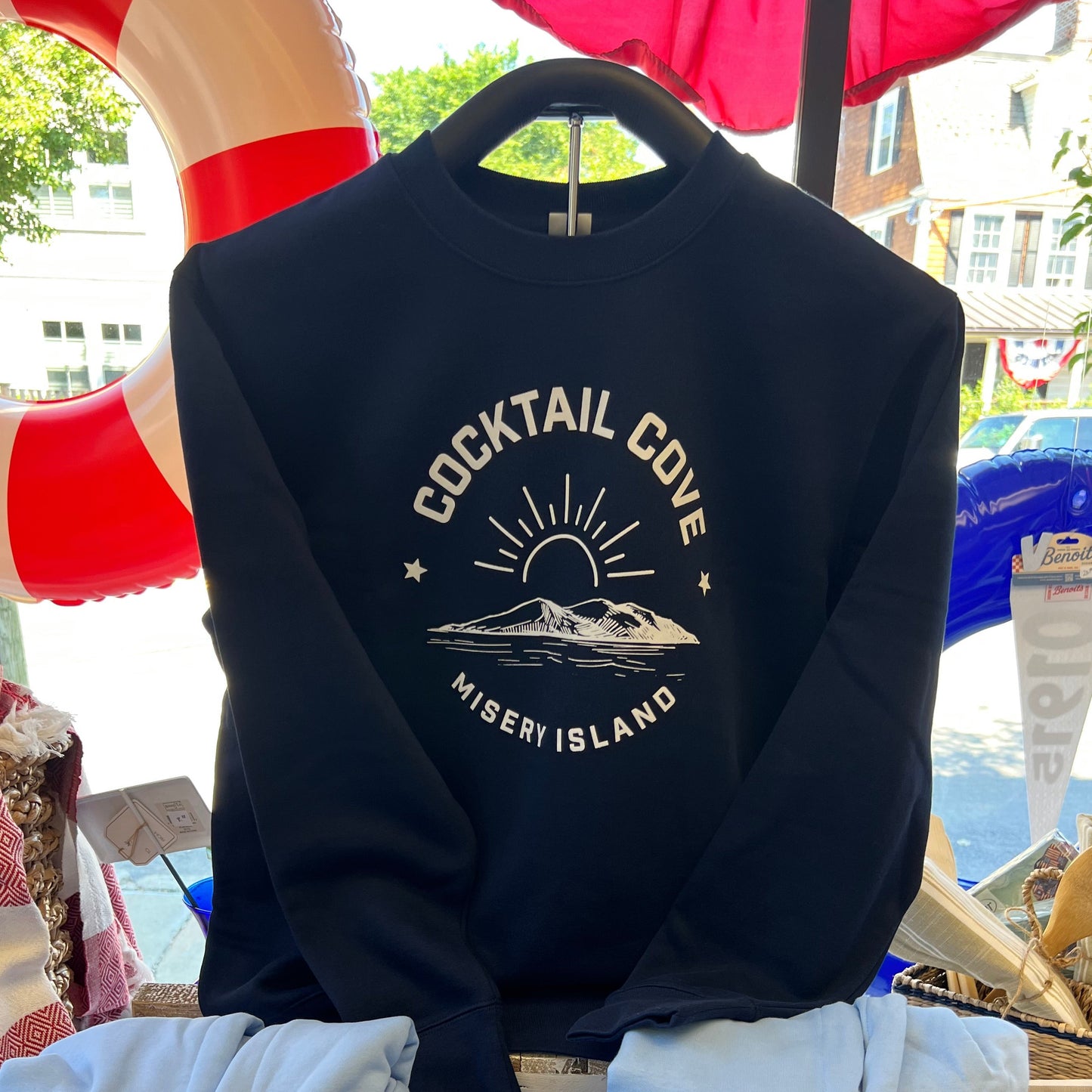 A navy crewneck sweatshirt hanging on a hanger, featuring a cream-colored graphic that reads 'COCKTAIL COVE' and 'MISERY ISLAND' with a sun and mountain illustration. The sweatshirt is displayed in a busy outdoor market setting with other items like light blue hoodies and red-and-white life preservers in the background.