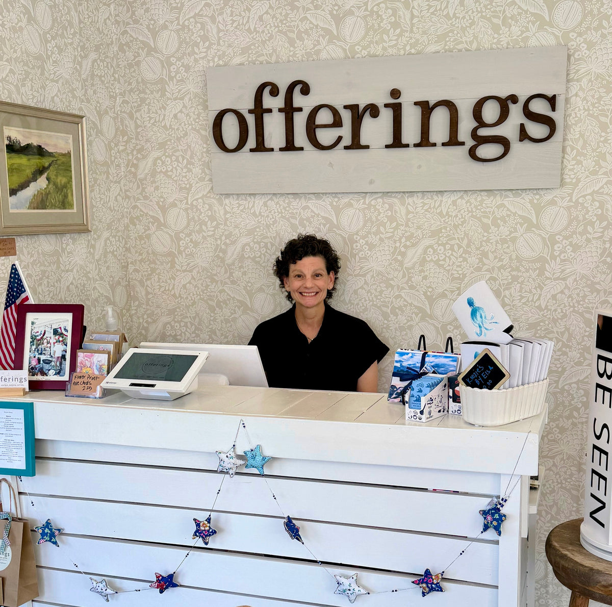 A smiling woman with short dark curly hair sits behind a white wooden counter in a shop or office. The background features a light beige floral wallpaper and a large wooden sign reading 'offerings'.