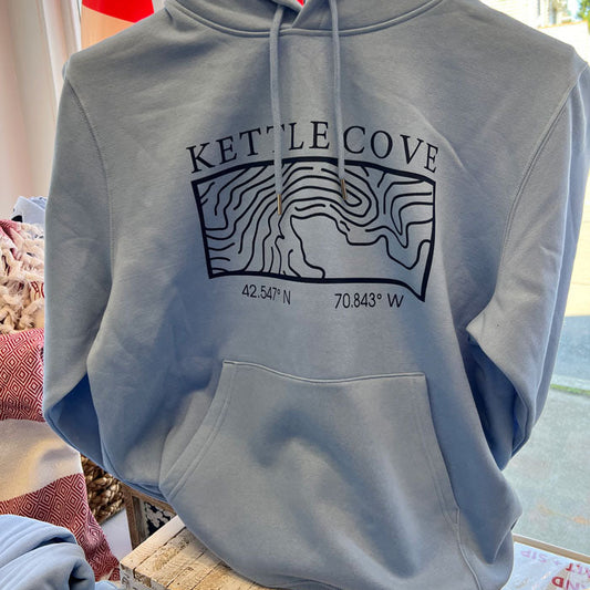 A light blue hooded sweatshirt hanging on a black hanger, featuring a black topographic map graphic with the text 'KETTLE COVE' and coordinates printed on the chest.