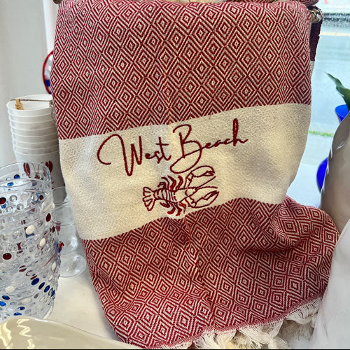 A woven beach towel or throw with a red and cream diamond pattern, featuring a cream horizontal band with 'West Beach' embroidered in red script and lobster illustrations. The towel is displayed on a table with a window and other items in the background.