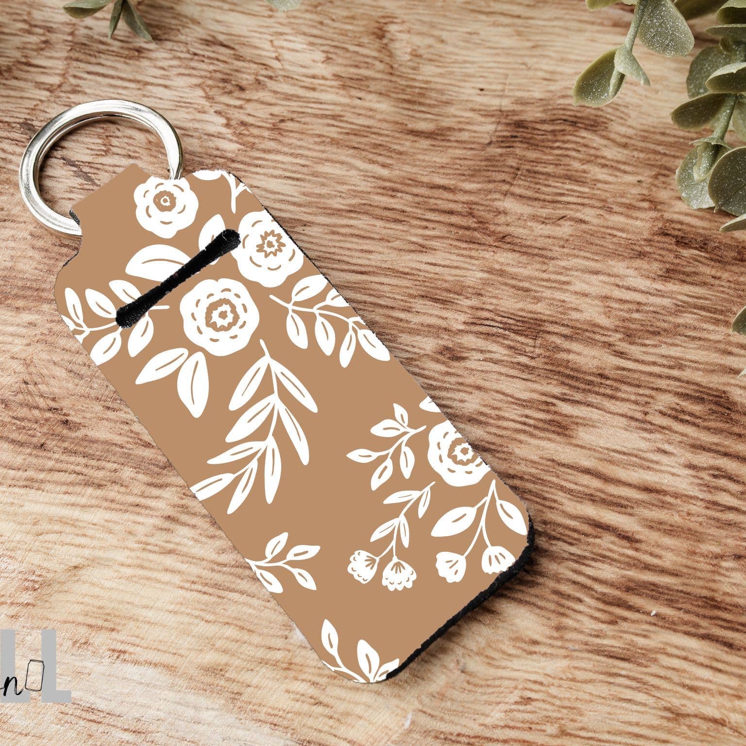 A rectangular keychain with rounded corners featuring a white floral and leaf pattern on a warm tan background, attached to a silver metal key ring. The keychain is photographed on a rustic wooden surface with small green leaves at the top corners.