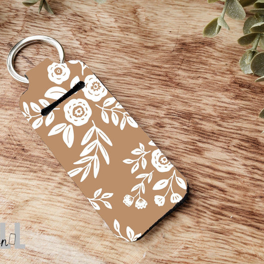 A rectangular keychain with rounded corners featuring a white floral and leaf pattern on a warm tan background, attached to a silver metal key ring. The keychain is photographed on a rustic wooden surface with small green leaves at the top corners.