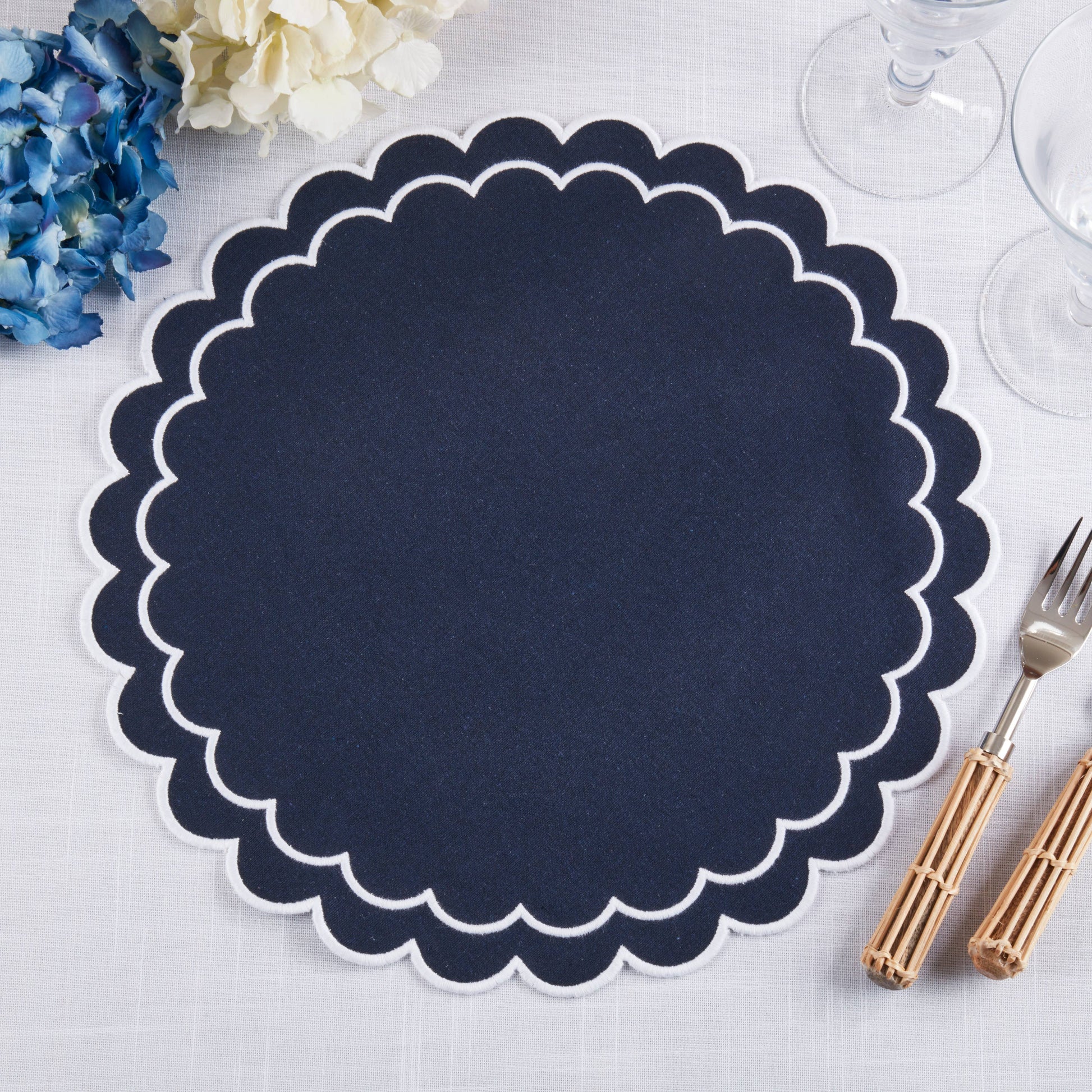 Navy blue scalloped placemat on a white surface with cutlery and flowers.