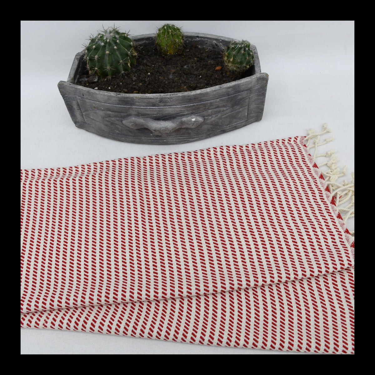 A folded red and white patterned Turkish cotton towel with cream fringe, displayed on a white background with a small gray wooden cactus planter behind it.