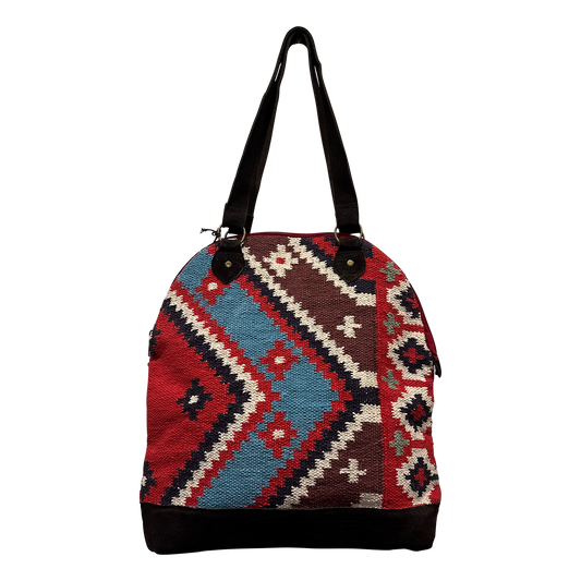 A woven shoulder bag with a geometric kilim pattern in red, blue, brown, and cream, featuring black straps and a black bottom base, shown against a plain white background.