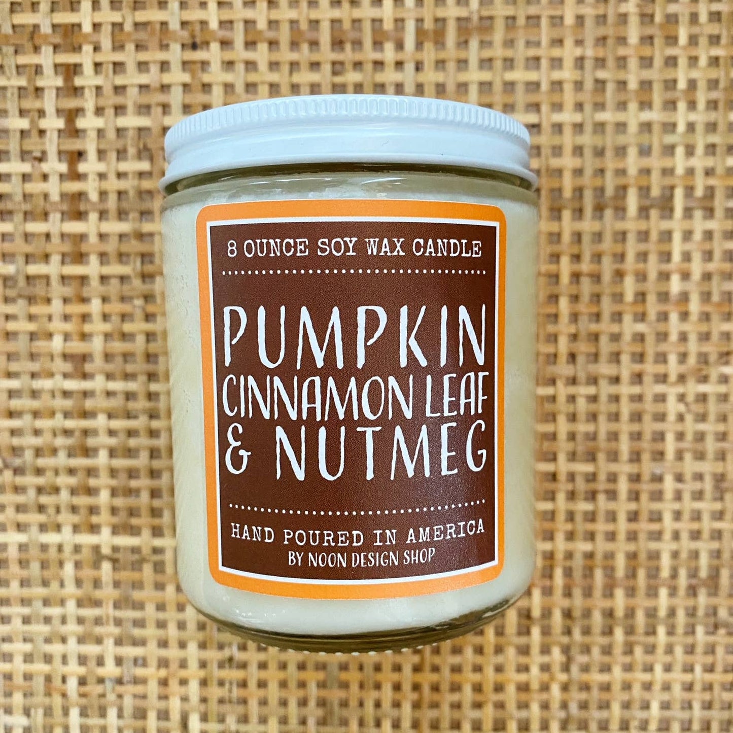 An 8-ounce soy wax candle in a clear glass jar with a white lid, featuring a brown label with orange borders that reads 'Pumpkin Cinnamon Leaf & Nutmeg' and 'Ncon Design Shop,' set against a woven tan background.