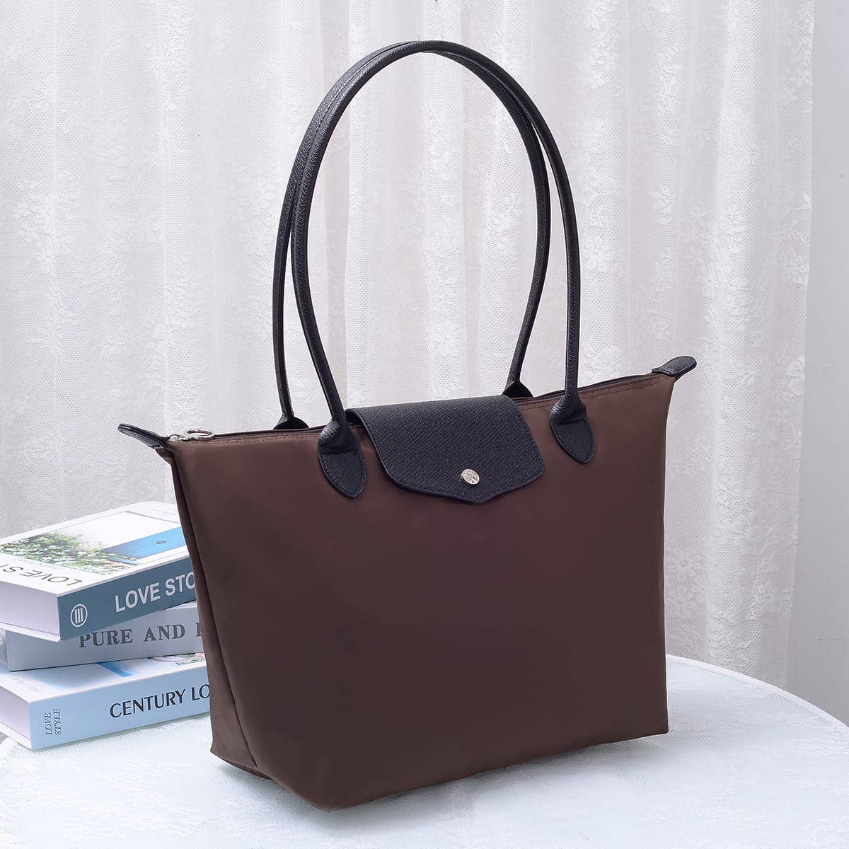 A brown and black leather tote bag with double shoulder straps, displayed on a white table next to a stack of books in front of a white curtain background.