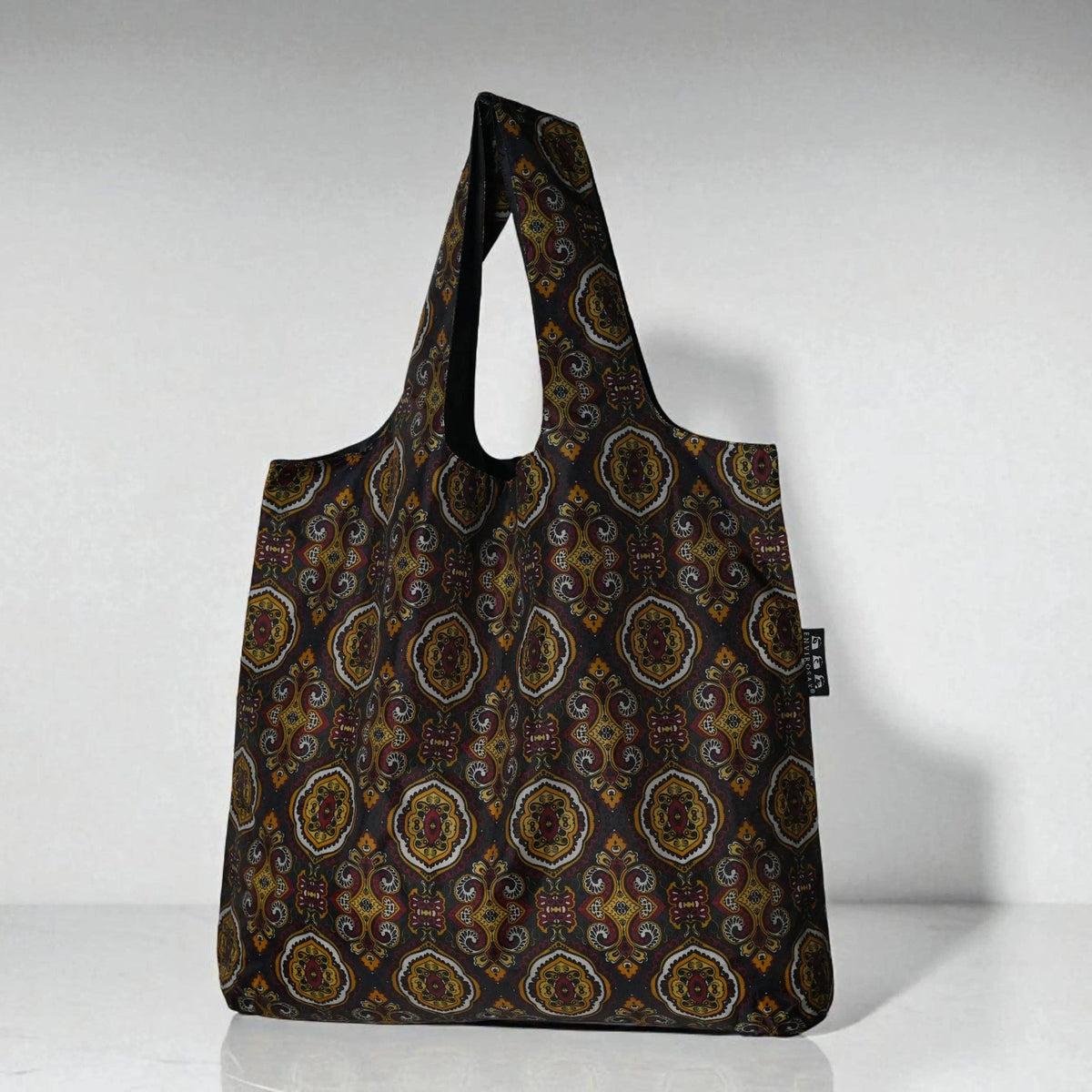 A reusable tote bag with a dark brown background and an intricate multicolor geometric medallion pattern, featuring two shoulder straps and a small black brand tag on the side.