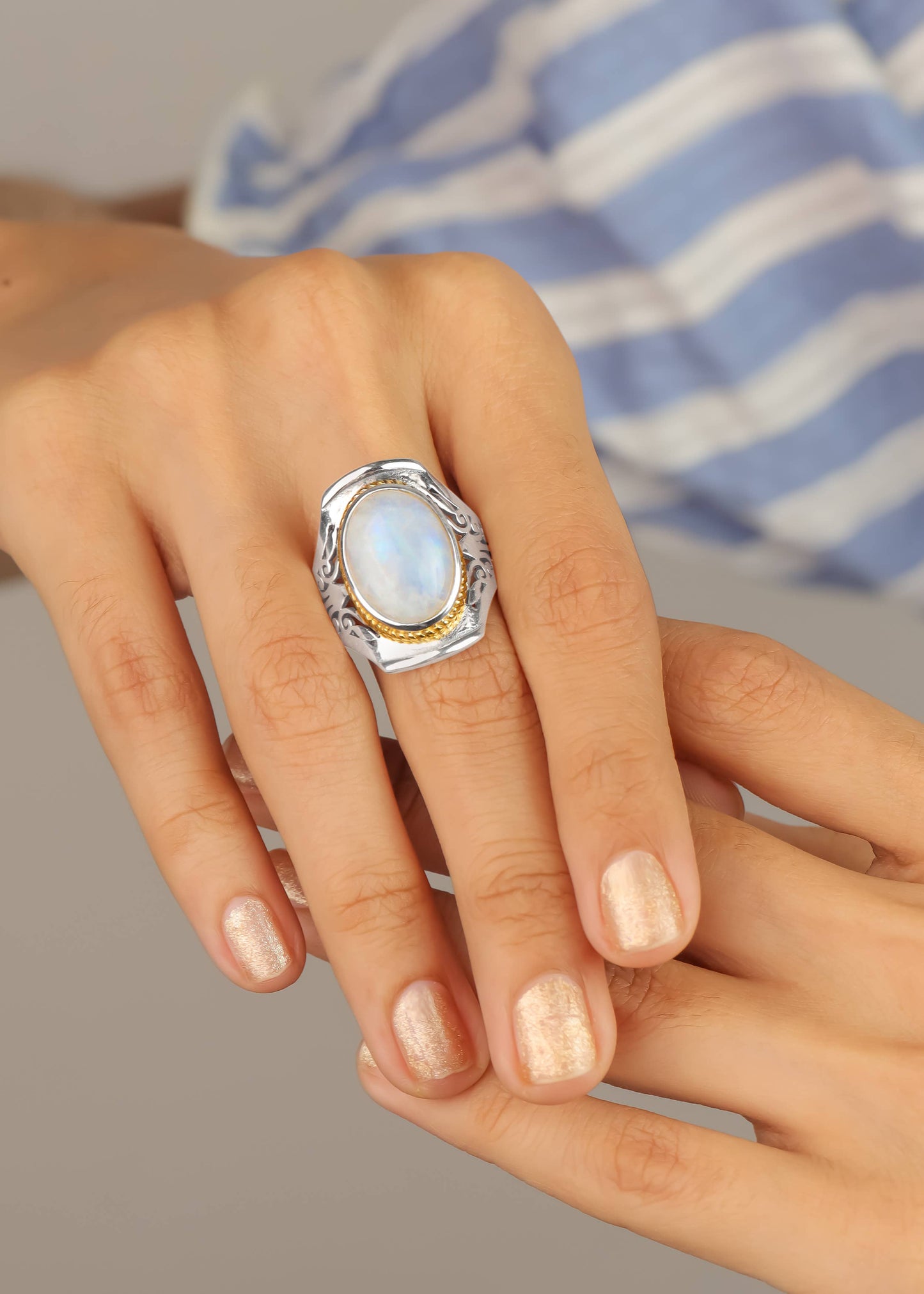 A close-up of a hand wearing a large silver ring with a white moonstone and a gold accent band, set against a soft-focus background of a blue and white striped shirt.