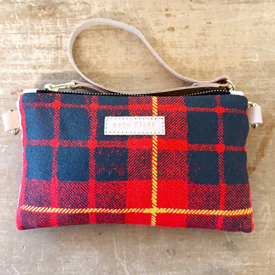 A small zip pouch with a red and navy tartan plaid pattern, featuring a light beige wrist strap and a white rectangular brand patch on the front, displayed on a rustic wooden surface.