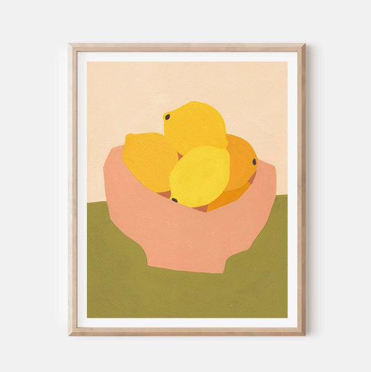 A framed minimalist art print depicting yellow lemons in a pink bowl against a beige and olive green background, mounted on a light gray wall.