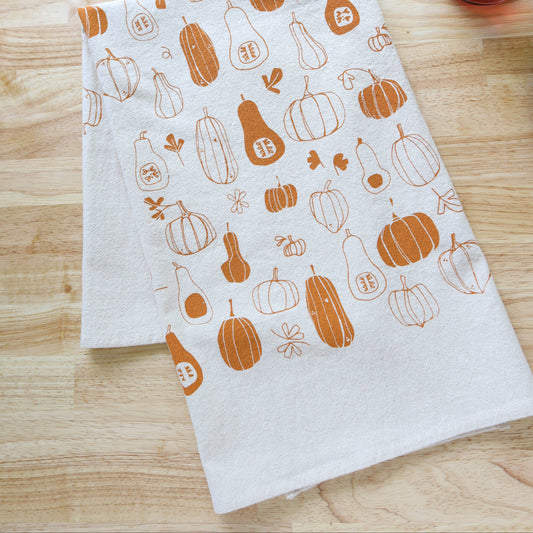 A folded white kitchen towel with an orange pumpkin and pear pattern, displayed on a light wood tabletop next to a glass of iced tea.