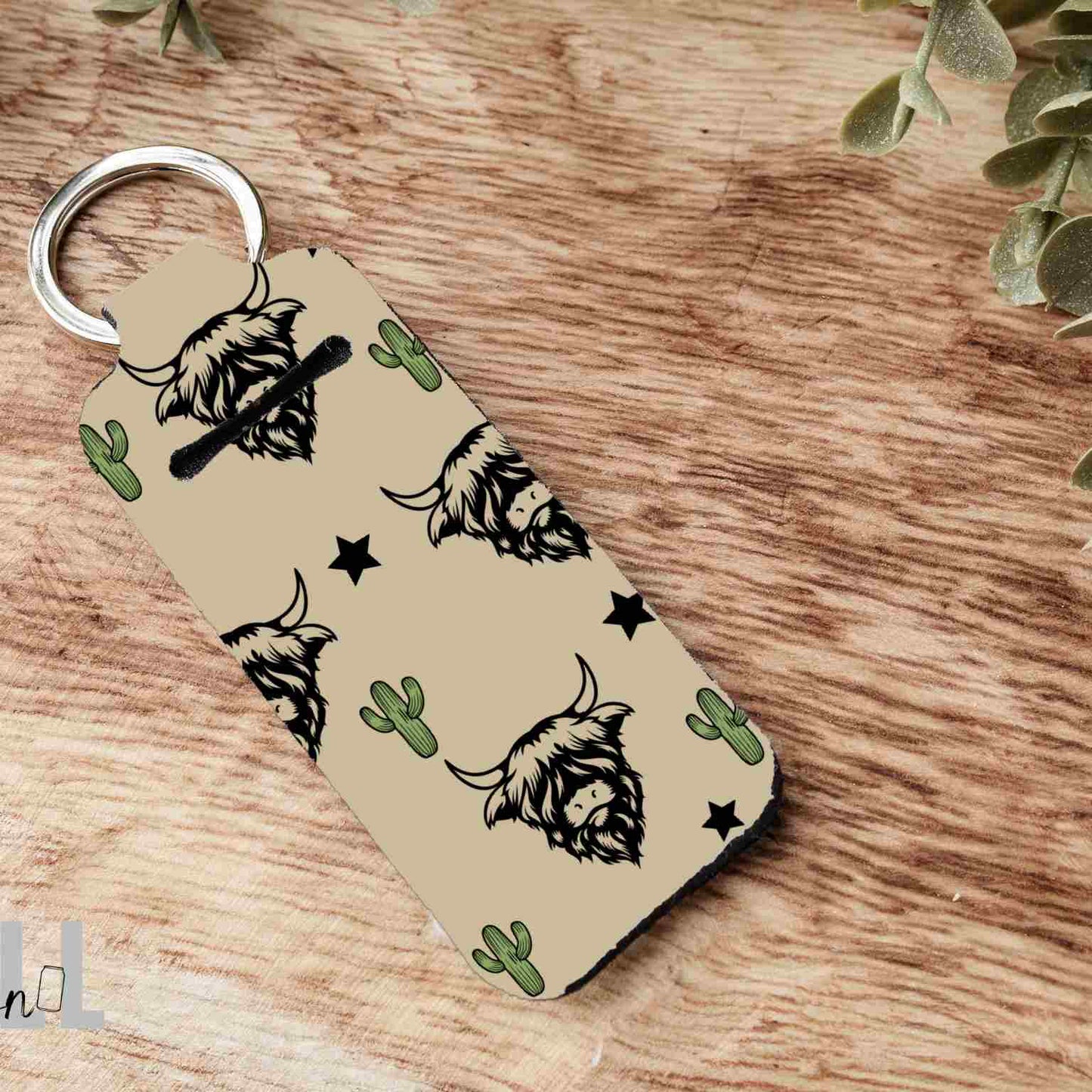A rectangular keychain with a silver split ring, featuring a tan background with black highland cow illustrations, green cacti, and black stars. The keychain is displayed on a rustic wooden surface with green leaves in the corners.