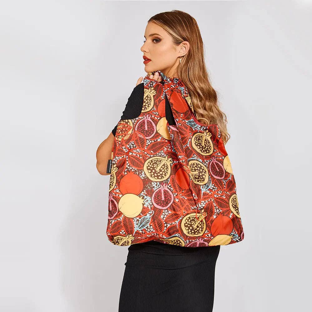 A woman wearing a black top and skirt models a red drawstring tote bag with a pomegranate print, shown from a side-back angle against a light gray studio background.