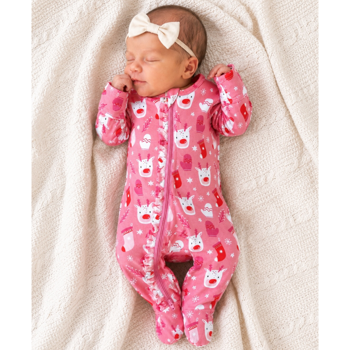 A sleeping newborn baby wearing a pink one-piece footed sleeper with a white and red holiday print, along with a cream bow headband, lying on a textured cream knit blanket.