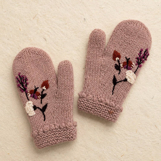 A pair of knitted fingerless gloves in dusty mauve pink, decorated with embroidered foxes and flowers on the back of the hands, displayed on a textured cream paper background.