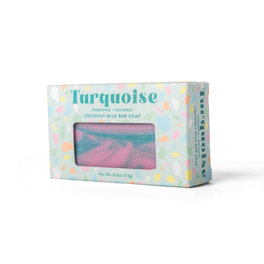 Turquoise branded coconut milk bar soap in a pastel patterned box with a clear window showing pink and blue marbled soap inside, photographed on a white background.